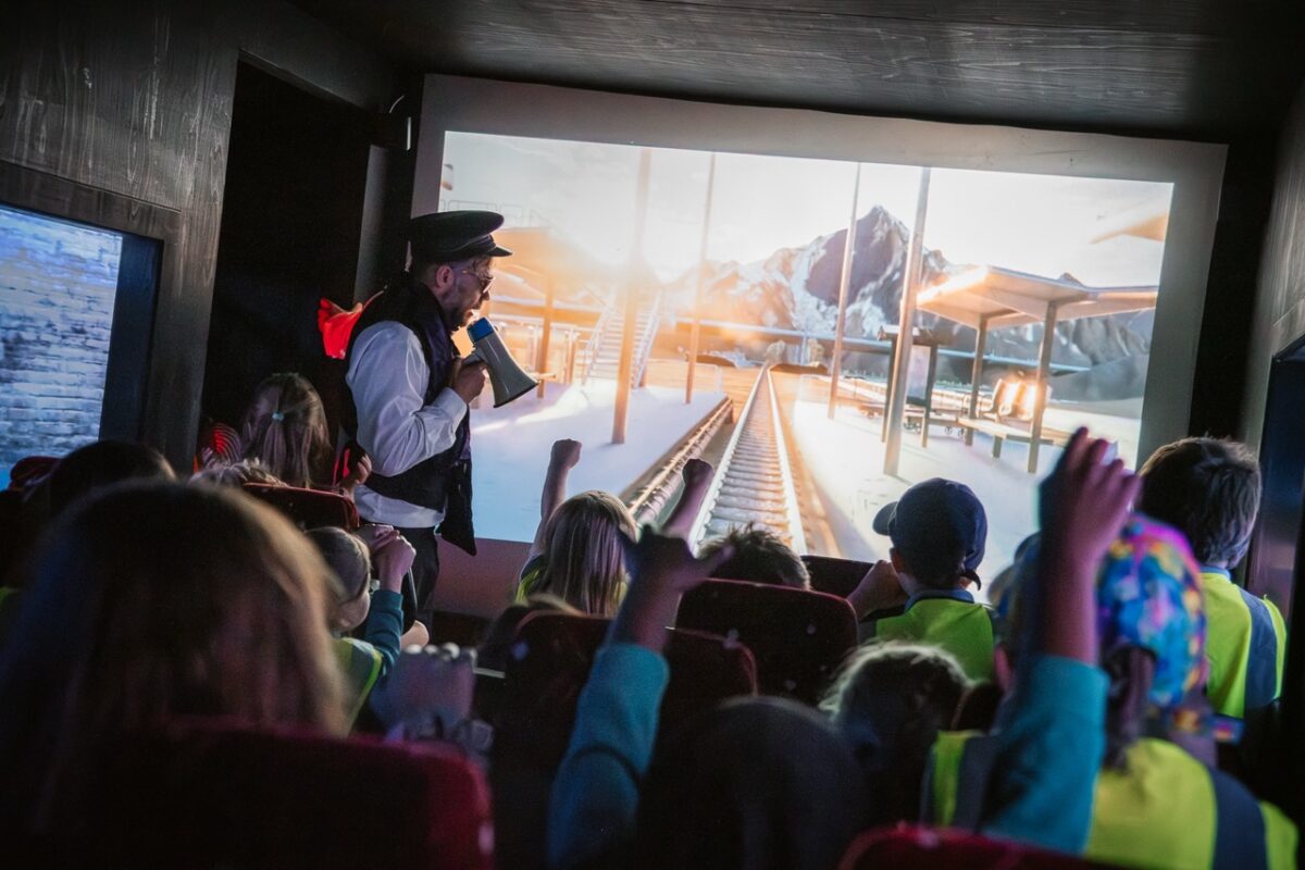 A photo of a performer talking into a megaphone in front of a projection of train tracks. there is a crowd of children watching the performance. 